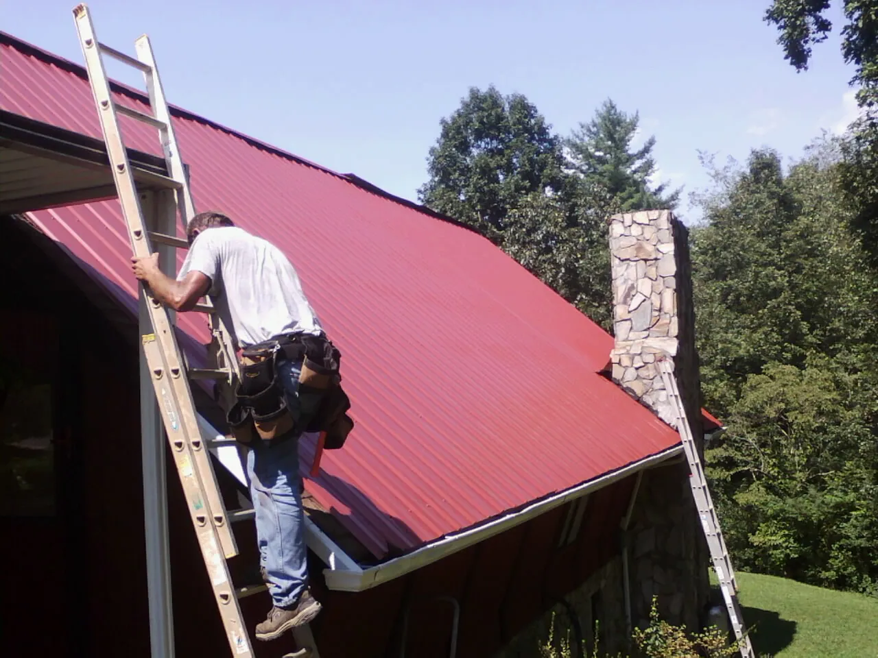Roofer on a ladder beside a red metal roof and stone chimney — Soffit & Fascia Repair by Roofing Brothers in Curtice